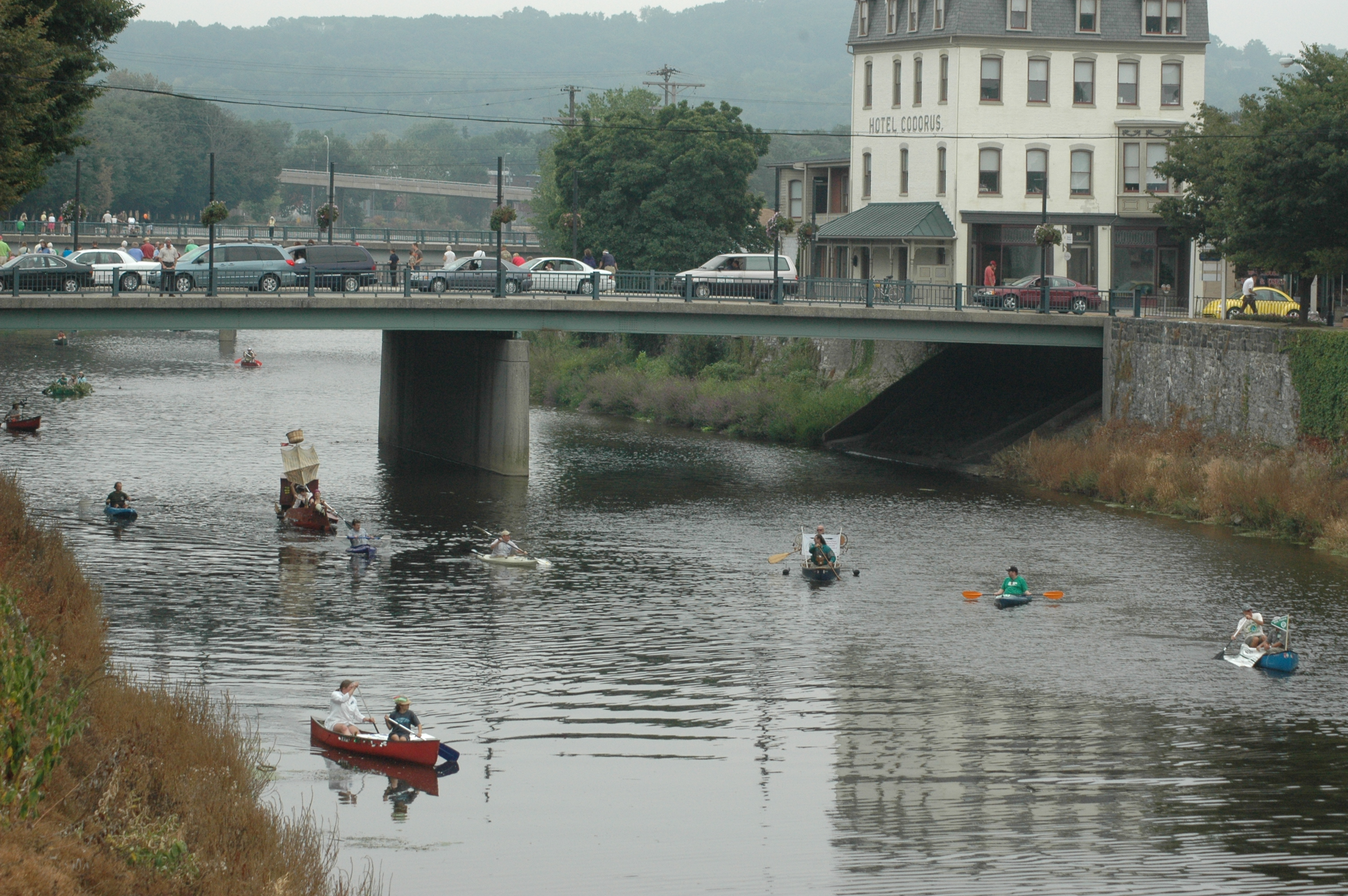 People Kayaking Down a River With Bridge Full of Cars in Background